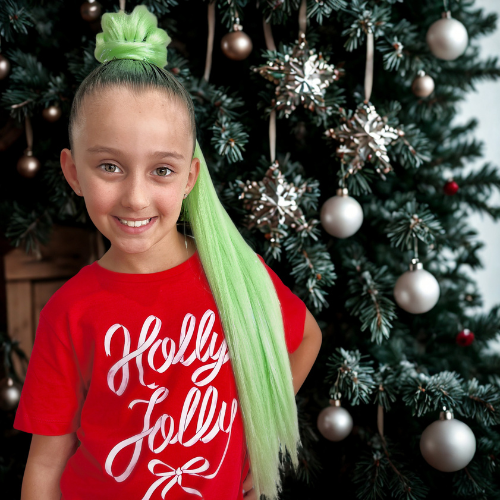 Child wearing a red 'Holly Jolly' shirt with green hair extensions in front of a decorated Christmas tree.