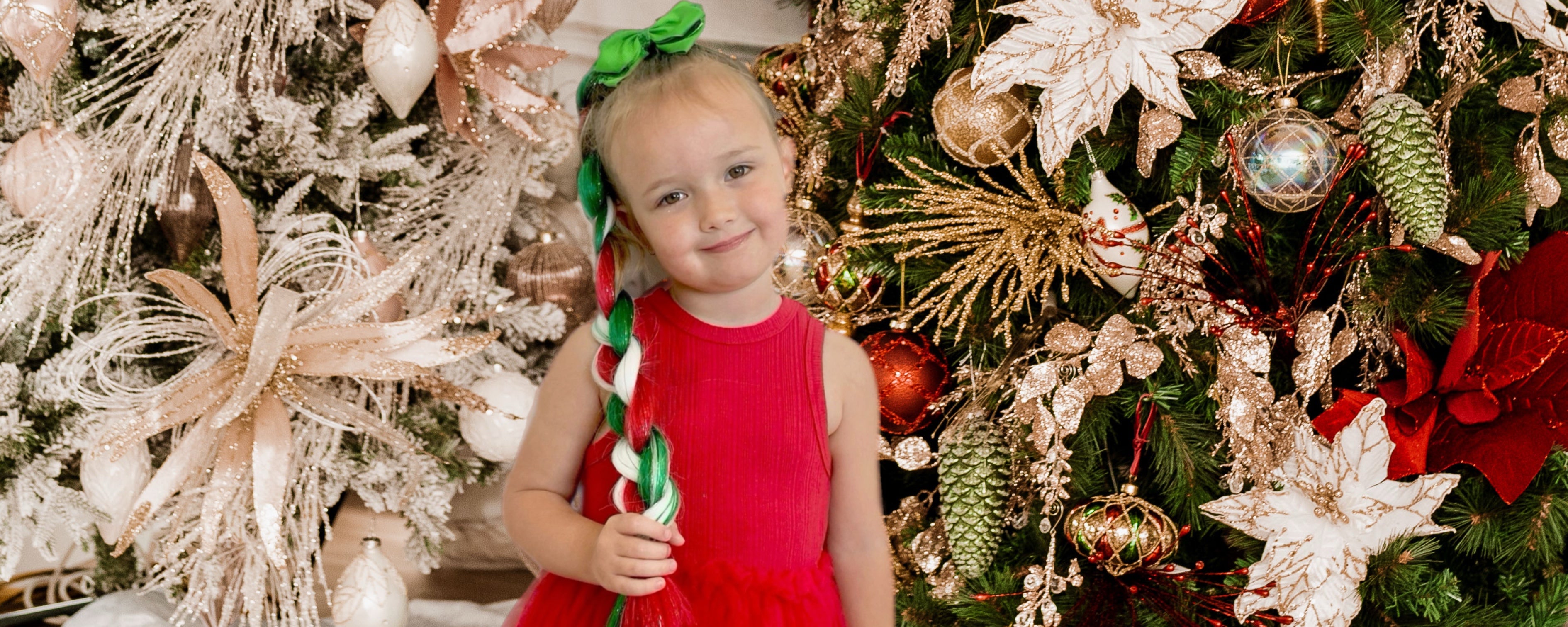 Child in a red dress wearing reusable mermaid ponytailstanding in front of Christmas tree.